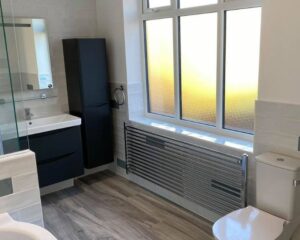 A new bathroom that has been fitted with blue cabinets and cupboards, wood-effect vinyl flooring, a new toilet and metal radiator.