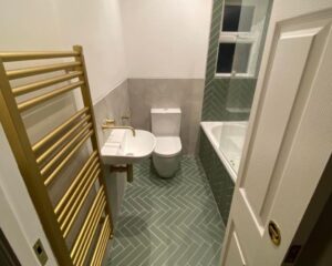 A new bathroom fitted with a gold towel rail, green herringbone tiled floors, a new floating sink, a new toilet and a bath over shower.