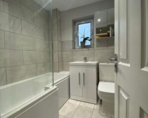 A new bathroom fitted with a new bath over shower, sink, toilet and grey tiled walls and floors.