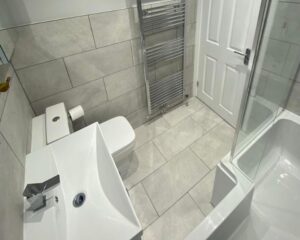 A new bathroom fitted with a new bath over shower, sink, toilet and grey tiled walls and floors.
