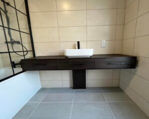 A bathroom featuring a floating wooden counter with a sink, cream wall tiles and grey floor tiles with a bathtub and shower.