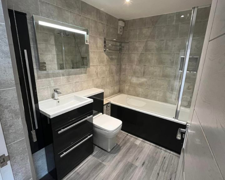 A new bathroom installation in a house in Southampton featuring grey wooden vinyl flooring, blue cabinets with sink, a bathtub with shower, a new toilet and grey wall tiles.