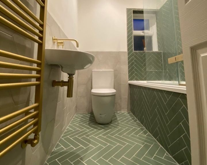 A new bathroom fitted with a gold towel rail, green herringbone tiled floors, a new floating sink, a new toilet and a bath over shower.